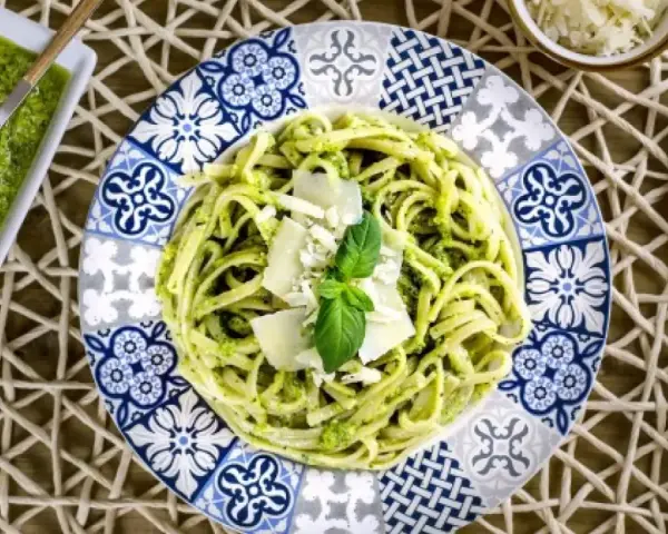 Pasta al pesto servida en plato decorado, con salsa de pesto y queso rallado al lado.