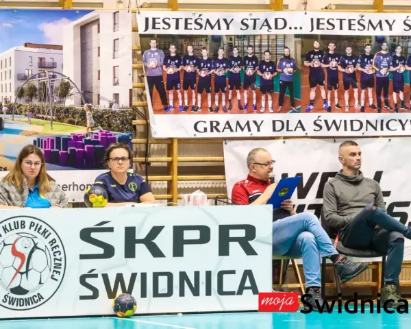 Na banerze widnieje drużyna SKPR Świdnica. Dwie kobiety siedzą za stołem z piłką ręczną.