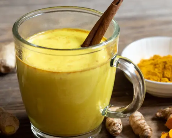 Taza de leche dorada con canela, rodeada de raíz de cúrcuma fresca y en polvo.