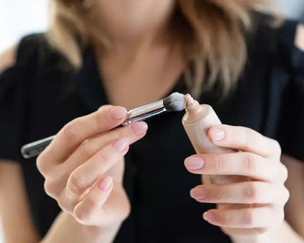 Dłonie z perfekcyjnym manicure nakładają pędzelkiem dr irena eris podkład rozświetlający na pociągłą tubkę.