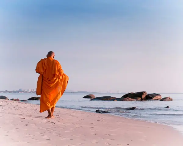 A modern day monk in an orange robe walks along a serene beach at sunrise, with calm ocean waves and distant city skyline.