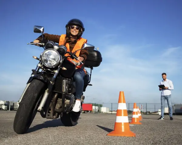 Joven sonriente en moto, lista para obtener el carnet de moto 49cc. Instructor observa en el fondo.