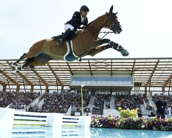 Un cavalier et son cheval s'envolent au-dessus d'une rivière lors d'un CSI Equitation. Le public applaudit dans les gradins.