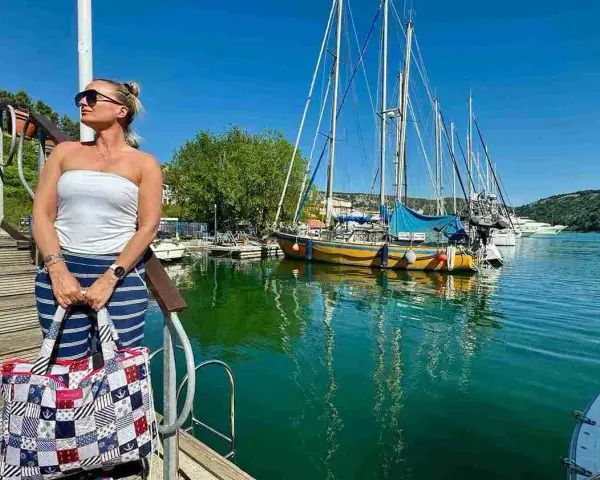 Kobieta z torbą na tle przystani z żaglówkami i turkusową wodą.