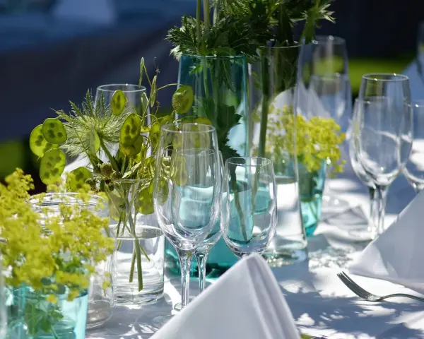 Eleganckie wesele w ogrodzie. Stół nakryty białym obrusem, ozdobiony zielonymi roślinami i kieliszkami.