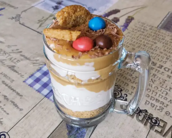Warstwowy deser banoffee w szklance, ozdobiony ciasteczkiem, cukierkami i wiórkami czekolady.