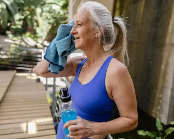 Femme d'âge mûr s'essuyant le visage avec une serviette bleue, tenant une bouteille d'eau. Elle fait du sport malgré la canicule.