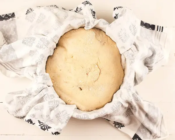 Masa de pan de patata cruda, cubierta con un paño blanco con estampado floral gris, lista para levar.