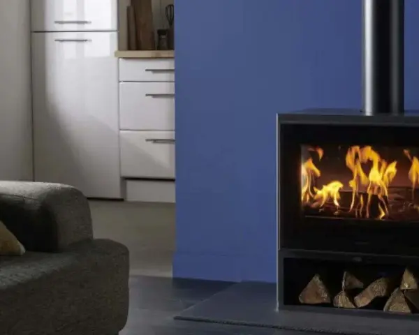 Estufa de leña moderna con fuego encendido y leña apilada, junto a un sofá y muebles de cocina.