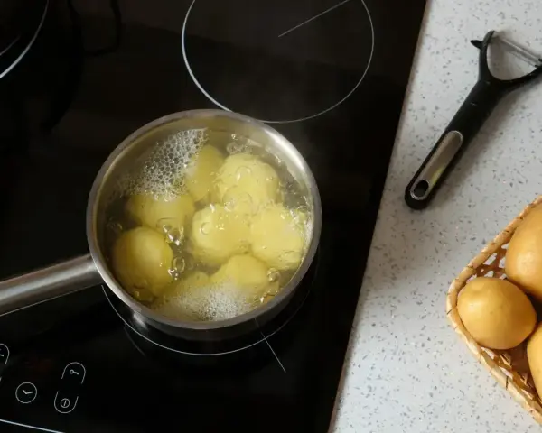 Ziemniaki gotują się w garnku na płycie indukcyjnej. Obok leży obieraczka i koszyk ze świeżymi ziemniakami. Oto jak ugotować ziemniaki.