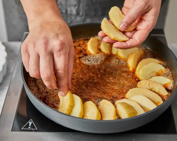 Hände legen Apfelschnitze in eine Pfanne mit karamellisierter Butter. Die Zubereitung der tarte tatin original beginnt.