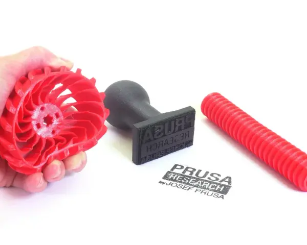Czerwony, elastyczny filament w kształcie wirnika, stempel z logo Prusa Research i czerwona rurka.