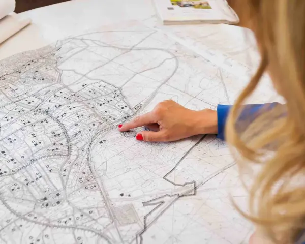 Kobieta z rudymi paznokciami wskazuje palcem na mapę. Plan miasta, z zaznaczonymi ulicami i budynkami, sugeruje analizę terenu pod kątem jego **użytku ba**.