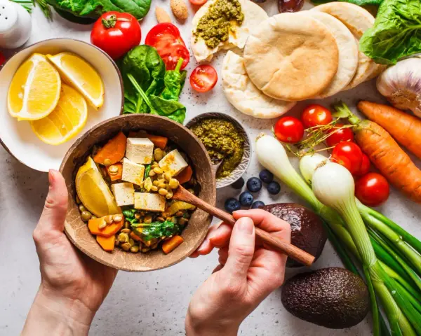 Zdrowa dieta na stawy: miska pełna soczewicy, tofu, warzyw i cytryny. Obok składniki na pyszne danie: pomidory, marchew, awokado, czosnek, cebula, jagody, pesto i chlebki pita.