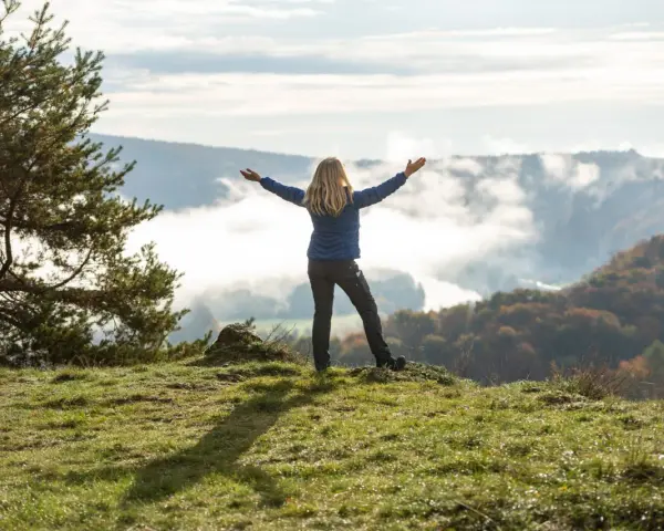 Frau mit ausgebreiteten Armen auf einem Hügel, umgeben von Nebel und Herbstfarben. Das Gefühl von Freiheit und die Bedeutung des Pilgerns werden hier spürbar.