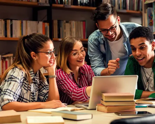 Vier Studierende lernen in der Bibliothek. Ein Mann, der wie ein seelsorger quereinsteiger wirkt, erklärt etwas am Laptop.