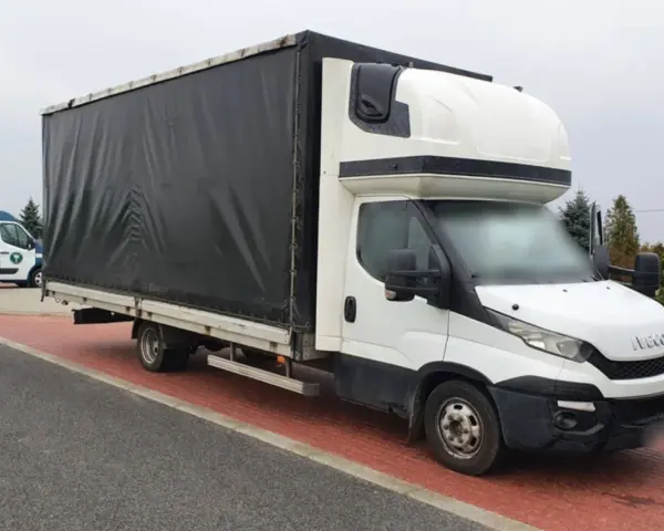Biały bus Iveco z czarnym plandeką, ładowność 1,5 tony, stoi na czerwonej kostce brukowej.