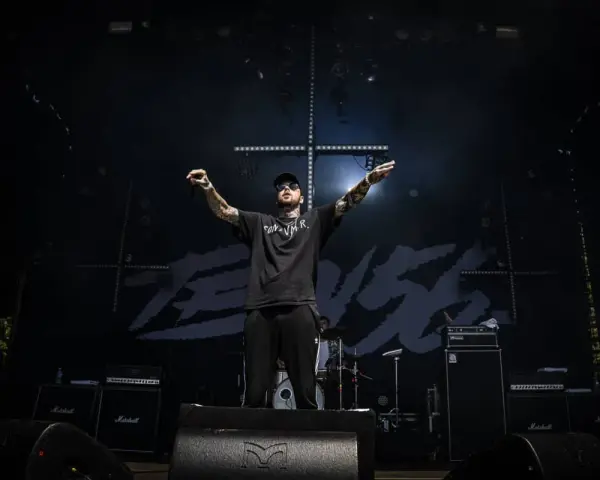 Un rappeur tatoué, casquette et lunettes de soleil, bras levés sur scène, devant un fond noir avec le logo "TEN 56".