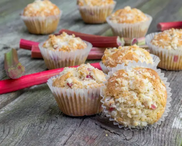 Pyszne muffinki z rabarbarem, posypane kruszonką, leżą na drewnianym blacie obok świeżych łodyg rabarbaru.