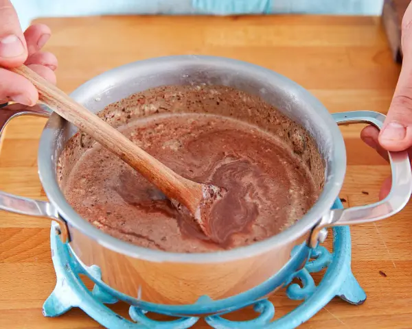 Hände rühren eine schokoladige Masse in einem Topf. So einfach lässt sich die Glasur selber machen für Kuchen oder Gebäck.