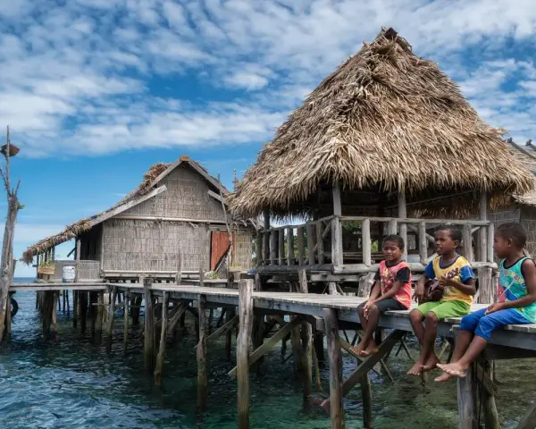 Trzech chłopców siedzi na drewnianym pomoście przy tradycyjnych chatach na wodzie w Raja Ampat.