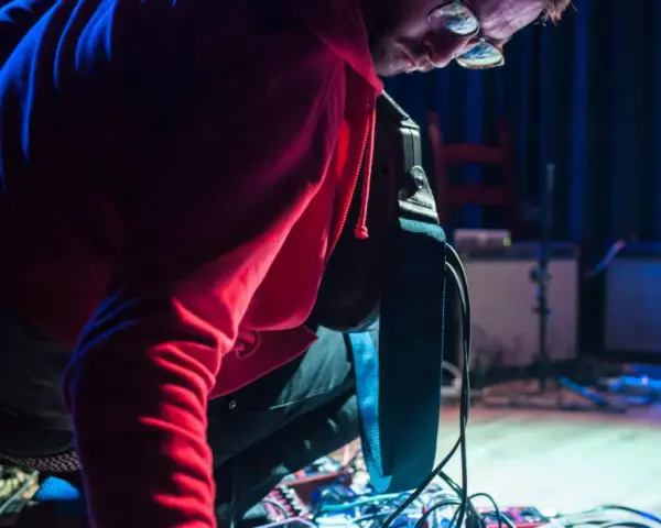 Musiker mit roten Kapuzenpullover beugt sich über ein Pedalboard, um den "Delay"-Effekt einzustellen.