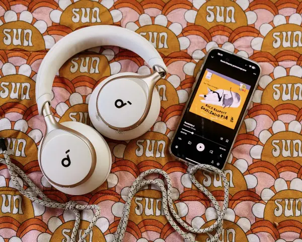 Białe słuchawki i smartfon z okładką audiobooka "Kocie kłopoty grzecznego psa" leżą na kolorowym tle z motywem słońc. Idealne do słuchania książek z lektorem.