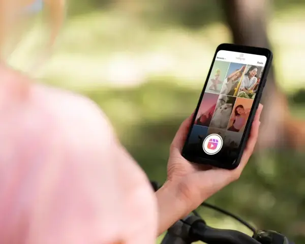 Osoba trzyma telefon z widokiem na ekran Instagrama, gdzie widać siatkę zdjęć i ikonę Reels. Dowiedz się, jak zrobić rolkę na Instagramie ze zdjęć.