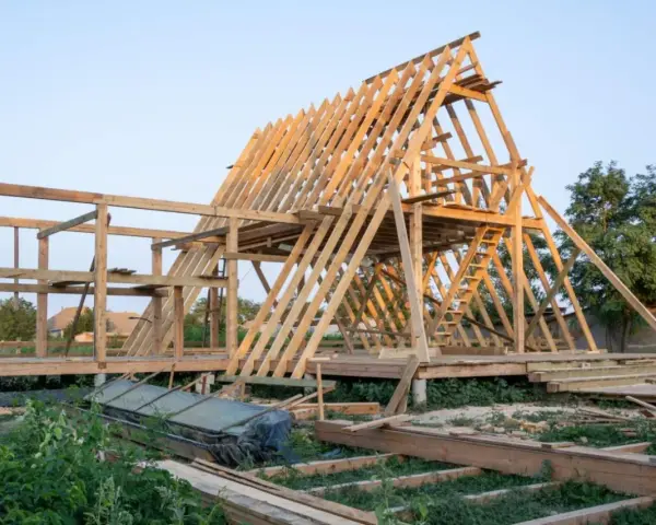 Drewniana konstrukcja domu w budowie. Widoczny szkielet dachu i ścian, schody. Fundament punktowy pod domek drewniany jest już gotowy.