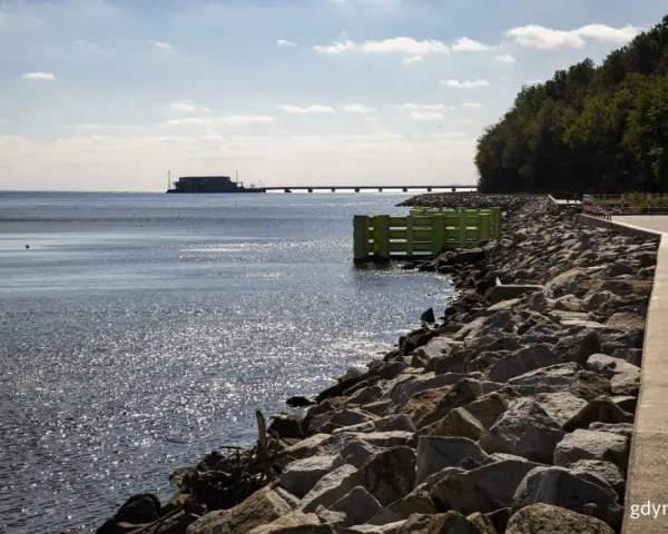 Kamieniste wybrzeże w Gdyni Oksywie z zielonym pomostem i falochronem. W oddali widać most i budynek na wodzie.