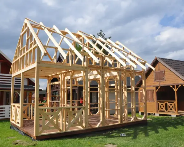 Drewniany projekt domu szkieletowego w budowie. Widać konstrukcję ścian i dachu, taras oraz balustrady.