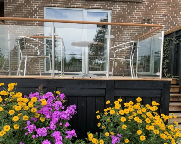 Vacker altan med glasräcke och trädetaljer. Blommor i gult och lila ramar in staketet. Perfekt altan staket inspiration för en modern uteplats.
