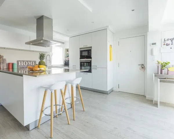 Moderna cocina blanca y gris claro con isla central, taburetes y frutas frescas.