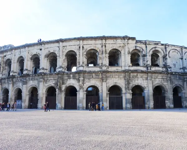 The ancient Roman amphitheater in Nîmes, France, stands majestically under a clear blue sky. Tourists gather in the plaza, ready to explore this historic marvel.