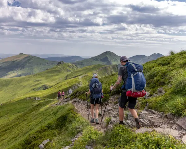 Randonnée sur le Puy Mary, des marcheurs explorent un sentier rocailleux bordé de verdure, avec des montagnes ondulantes en arrière-plan sous un ciel nuageux.