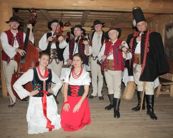 Zespół góralski w strojach ludowych, z instrumentami. Wśród nich skrzypkowie, basista i panie w tradycyjnych sukniach.
