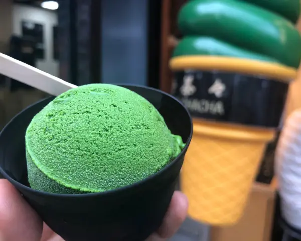 Zielone, japońskie lody matcha w czarnym kubeczku, z drewnianą łyżeczką. W tle wielki, żółty rożek z lodami.