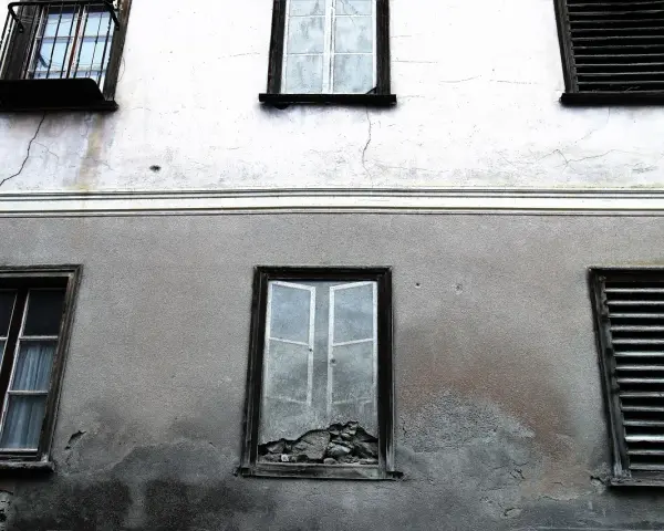 Stara elewacja z widocznymi pęknięciami i zaciekami. Okna z żaluzjami i jedno wybite.