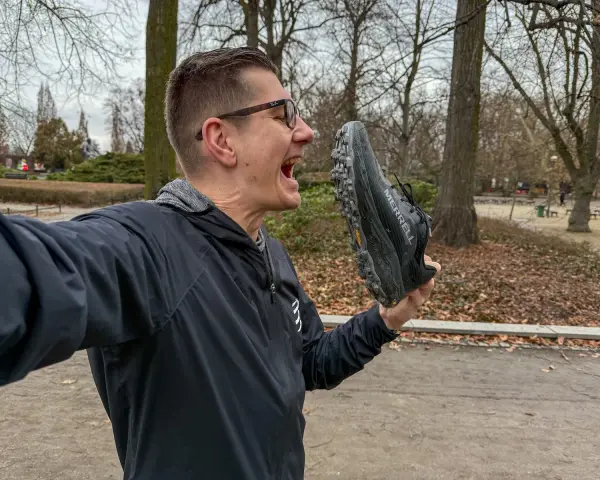 Mężczyzna w okularach trzyma but Merrell przed otwartymi ustami w parku.