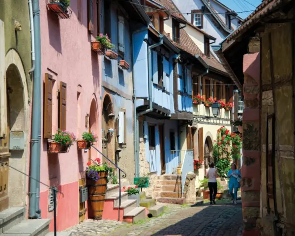 Charming street with colorful houses, perfect for day trips from Strasbourg. Two women stroll past flower-filled window boxes.