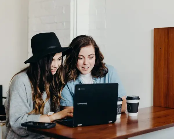 Dwie młode kobiety pracują na laptopie, jedna w kapeluszu, druga w dżinsowej koszuli. Ich wspólny projekt to passe recent.