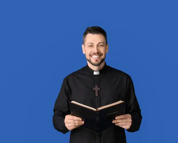Uśmiechnięty ksiądz w czarnej koszuli z krzyżem na szyi trzyma otwartą Biblię. W jego oczach widać ciepło, a na twarzy szczere słowa podziękowania.