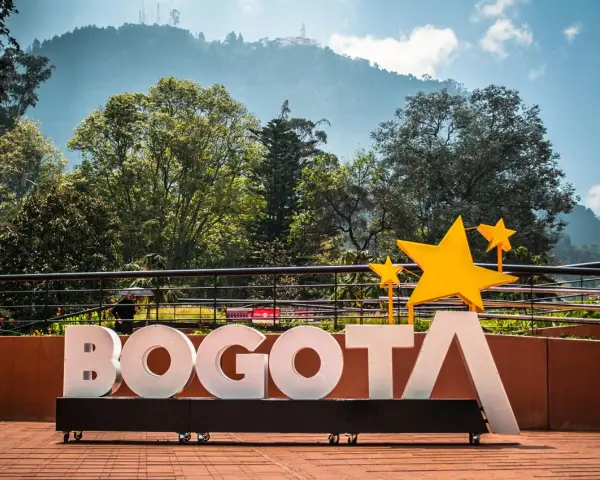Napis BOGOTA z żółtymi gwiazdami, symbolizujący stolicę Kolumbii, na tle zielonych wzgórz i błękitnego nieba.