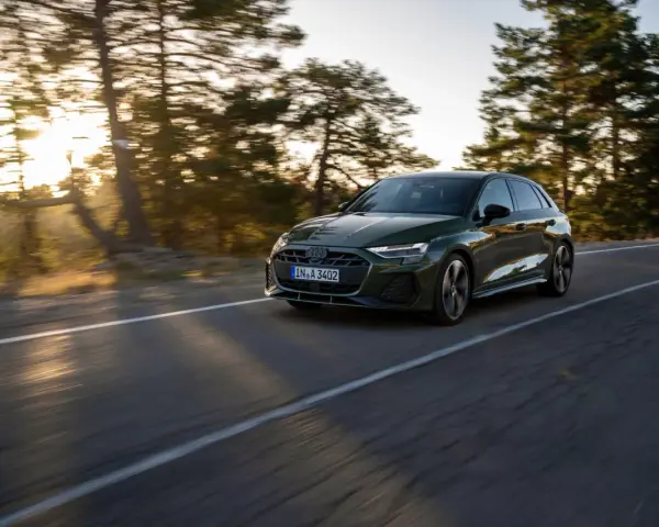 Un'auto verde scuro, un segmento premium, sfreccia su una strada panoramica al tramonto, con alberi sfocati sullo sfondo.