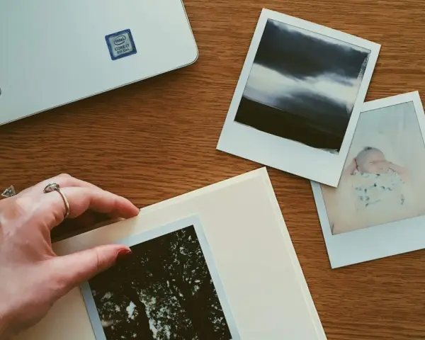 Dłoń układa zdjęcie na albumie, obok inne fotografie i laptop. Idealne miejsce, gdzie przechowywać zdjęcia z telefonu.