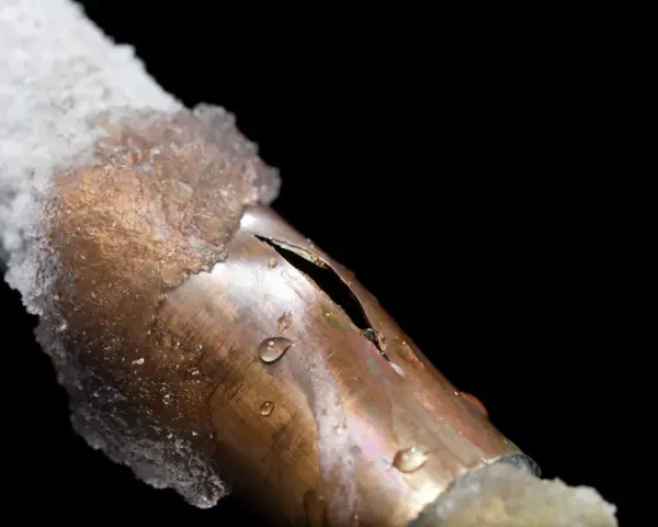 A copper pipe, partially covered in ice, shows a split from a frozen pipe repair. Water droplets cling to the damaged metal.