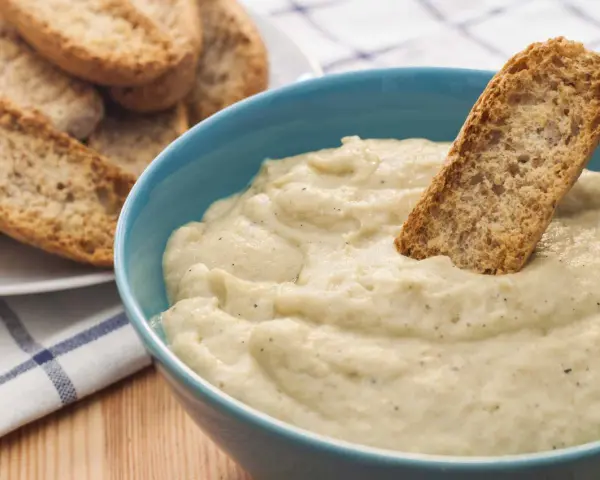 Un trozo de pan crujiente se sumerge en un cremoso baba ganoush en un cuenco azul. A un lado, hay una pila de pan tostado.