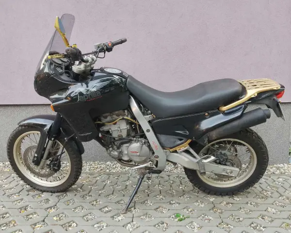 Czarny motocykl Aprilia Pegaso 650 z złotym bagażnikiem i lusterkami, zaparkowany na kostce brukowej.