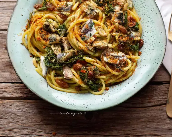 Un piatto di pasta con le sarde, condita con erbe aromatiche, pinoli e pangrattato tostato, servita su un piatto azzurro.