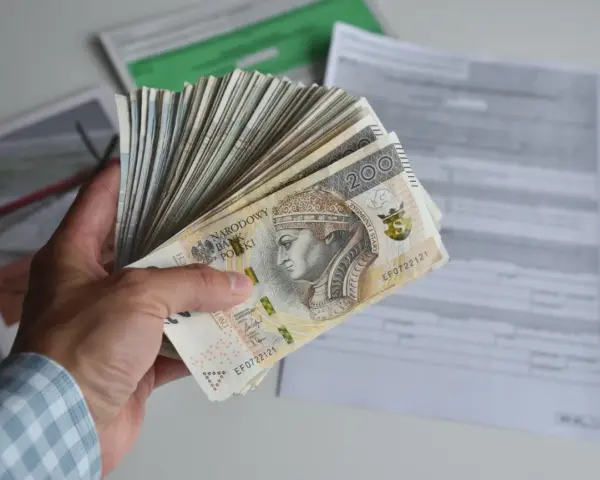 Dłoń trzyma plik banknotów 200 zł. W tle dokumenty, być może dotyczące tego, ile godzin to 0.5 etatu.